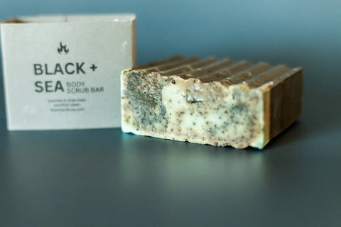 The Shanti + Tula Exfoliating Oatmeal & Shea Butter Body Scrub Bar sits before its packaging on a blue surface, showcasing its handcrafted, marbled texture with visible exfoliating bits and uneven edges.