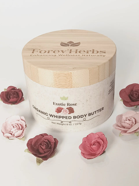 A jar of Forev'Herbs Exotic Rose Organic Whipped Body Butter with a wooden lid sits amid artificial pink and red roses on a white background. The label features rose graphics and natural wellness branding, emphasizing its nourishing formula.