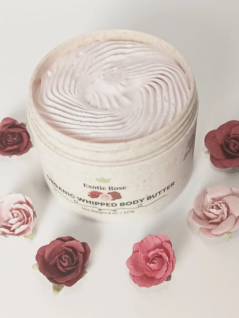 An open jar of Forev'Herbs Exotic Rose Organic Whipped Body Butter, featuring a swirl top, sits on a white surface surrounded by pink and red artificial roses.
