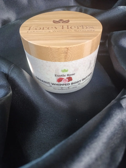 A jar of Forev'Herbs Exotic Rose Organic Whipped Body Butter with a bamboo lid rests on black satin, its label showcasing natural ingredients and wellness benefits.