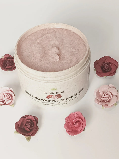 A jar of Forev'Herbs Exotic Rose Organic Whipped Sugar Scrub, featuring a light pink, fluffy texture, sits among artificial pink and red roses on a white background to showcase this natural exfoliating scrub.