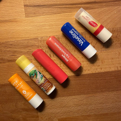 Six lip balm tubes in orange, yellow, green, red, blue, and white are displayed diagonally on wood—ideal for storing in the Tallac House Lip Balm Keychain Xtra-Large Lip Balm Holder (USA made, stainless steel) for larger diameter balms.
