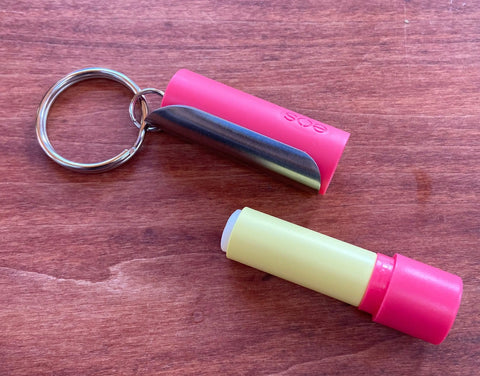 A Tallac House Lip Balm Keychain Xtra-Large Holder (stainless steel, USA made) is open on a wooden surface, showing its yellow lip balm stick and cap. Note: Fits larger diameter lip balms only.
