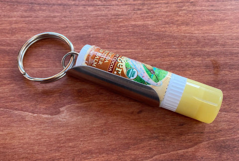 The Tallac House Lip Balm Keychain Xtra-Large Holder, made in the USA from stainless steel and designed for larger diameter lip balms, is shown resting on wood for convenient access.