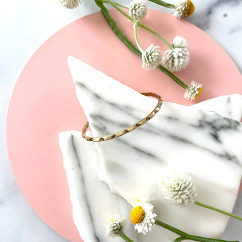 The GOLD CANAL BRACELET by Sarah Cecelia is displayed on white marble slabs atop a round pink surface, accented with delicate white and yellow flowers.