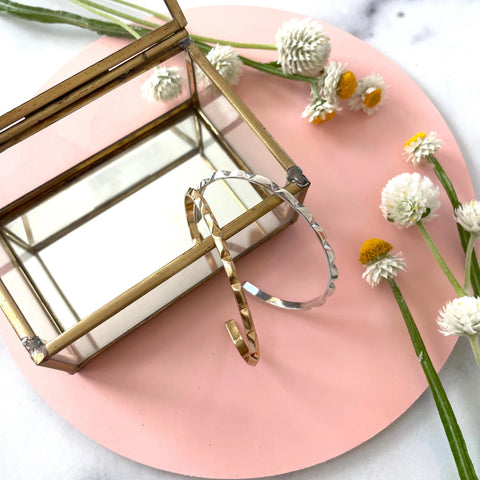The GOLD CANAL BRACELET by Sarah Cecelia rests on a glass and brass jewelry box atop a pink mat with scattered daisies—an elegant, American-made accessory.