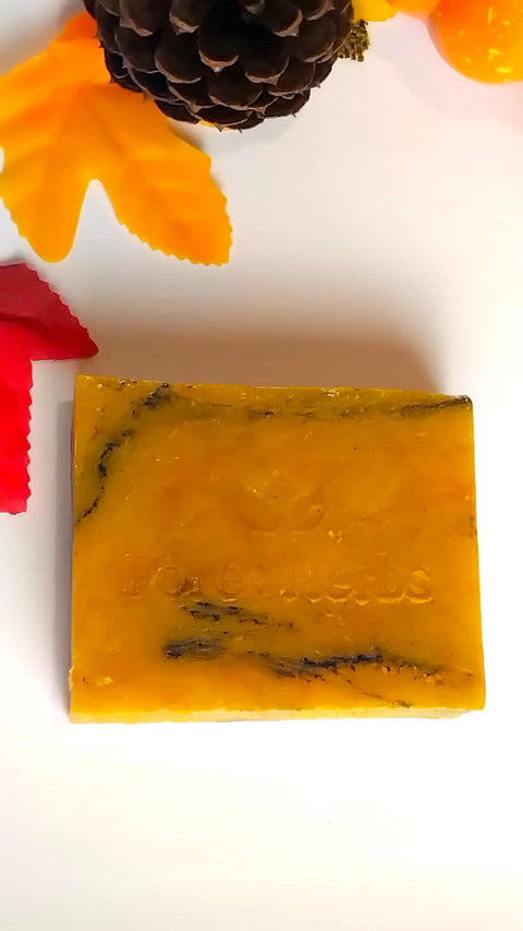 A rectangular yellow-orange soap with black swirls, Fall Festivities Handmade Soap by Forev'Herbs, rests on a white surface with autumn leaves and a pine cone—ideal for cozy fall collections and seasonal gifts.
