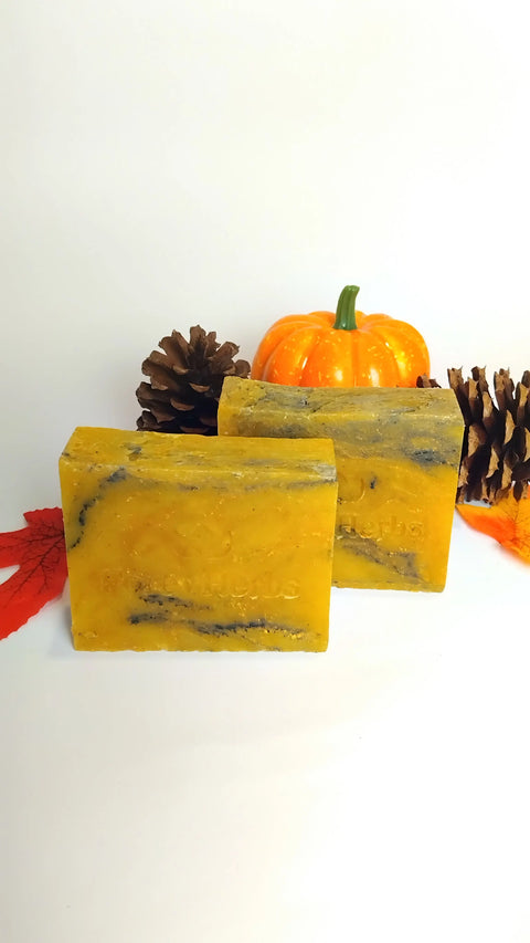 Two yellow-orange Forev'Herbs Fall Festivities Handmade Soap bars with black swirls are shown on a white background, accompanied by a small decorative pumpkin, pinecones, and orange leaves.