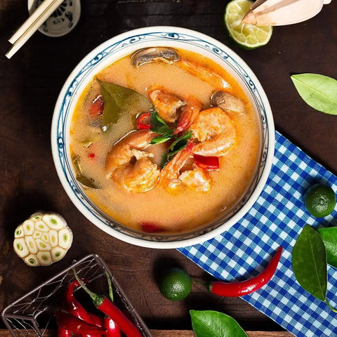 A bowl from vendor-unknown's Fancy Proteins & Truffled Sides | 4 Pack sits on a blue-checked cloth, garnished with herbs, red chilies, kaffir lime leaves, lime halves, and gourmet seasoning on a wooden table.