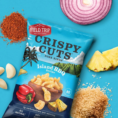 A pack of Field Trip Island BBQ 1oz (6 Pack) pork rinds is surrounded by sliced red onion, pineapple, brown sugar, bell pepper, garlic cloves, and BBQ seasoning—a tasty keto-friendly pineapple snack on a blue background.