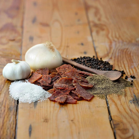 Field Trip Original Beef Jerky 12oz is displayed on a wooden surface with coarse salt, ground pepper, black peppercorns, a wooden spoon, and two onions—one partly peeled.