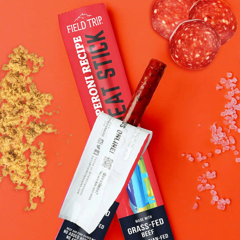 A partially opened Field Trip Pepperoni Recipe Meat Stick .5oz (24ct) is shown with pepperoni slices, pink salt crystals, and brown seasoning on an orange background.