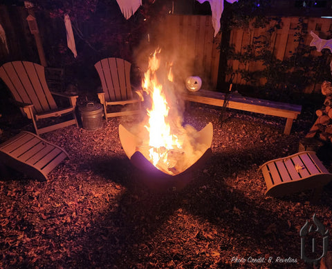 A Fire Flower Fire Bowl With Hollow Base by Ohio Flame burns brightly, surrounded by five wooden chairs. Halloween decorations and a pumpkin rest on a bench nearby in a backyard at night. Made in the USA.
