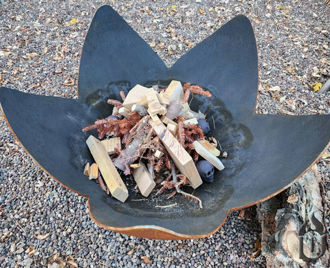 The Fire Flower Fire Bowl with Standard Base by Ohio Flame, a large black fire bowl shaped like a flower, stands amid pebbles and autumn leaves, filled with kindling and wood, ready for a cozy fire. Made in the USA.