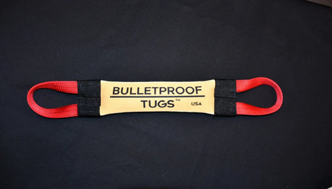 The Tooth Hugger Fire Hose Training Tug by Bulletproof Pet Products Inc features yellow and black USA-made fire hose, red handles, and BULLETPROOF TUGS USA in black text at the center. The toy is shown on a black surface.