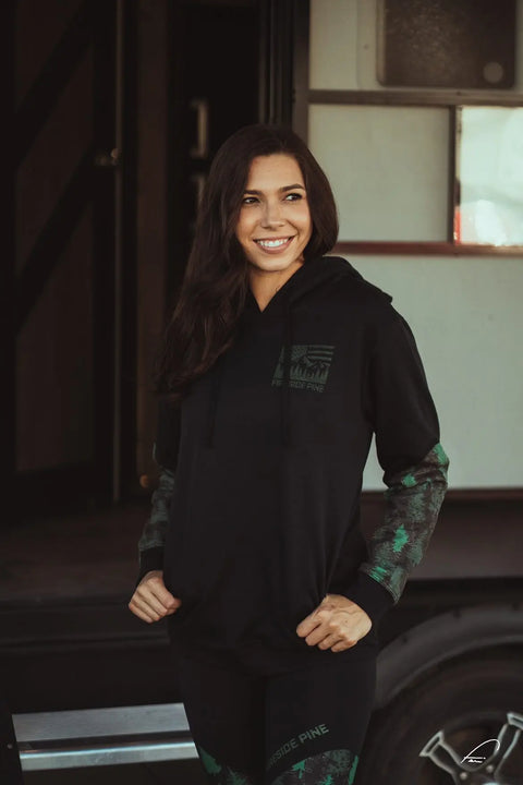 A woman with long dark hair, dressed in the Fireside Pine Black Cottonwood- Forest Green hoodie and black-green patterned leggings, stands smiling by an open trailer door, looking warm and cozy.