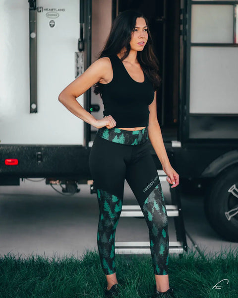 A woman in a black sleeveless top and Fireside Pine Aspen - Forest Green leggings stands on grass before a trailer, glancing aside with one hand on her hip.