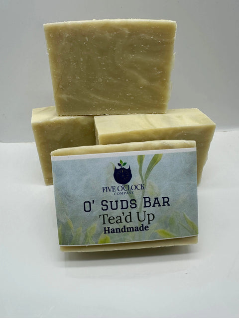 Three beige O’ Suds Bar soap bars from Five O Clock Co. are stacked, with one in front showing its label, set against a light blue and green leafy background.