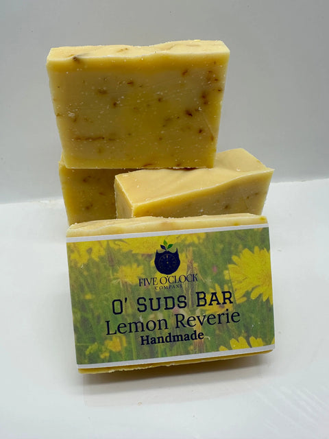 Three small-batch O’ Suds Bars by Five O Clock Co. are stacked, with the front bar showing its label and set against a backdrop of yellow flowers.