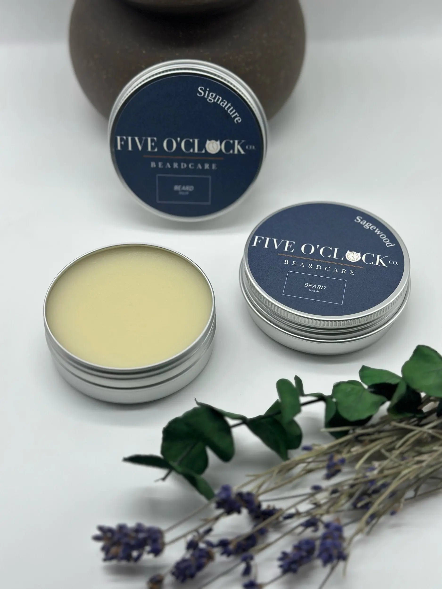 Two open tins of Five O Clock Co. Beard Balm Butter, one with the lid off revealing the balm, rest on a white surface beside dried lavender and a brown vase—a perfect scene for premium beard conditioning.