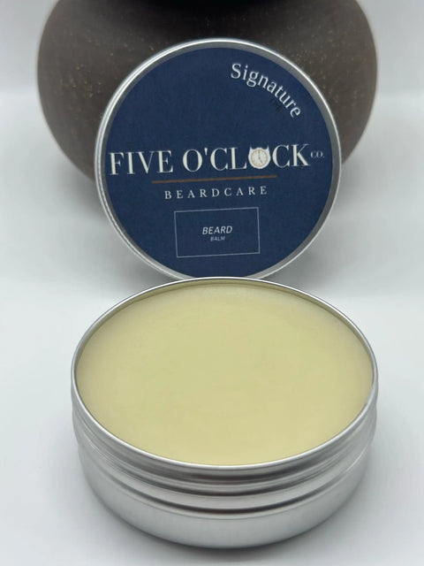 An open tin of Five O Clock Co. Beard Balm Butter shows the smooth balm inside, ready to nourish your beard. The dark blue lid with white text sits behind the tin against a softly blurred brown background.
