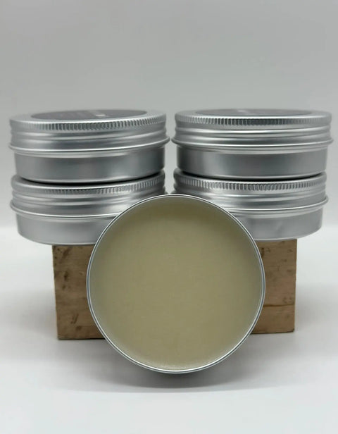 A round, open tin of Five O Clock Co. Beard Balm Butter sits before six closed tins stacked on a wooden block against a white background. Ideal for nourishing and conditioning your beard.
