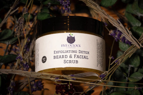 A jar of Five O Clock Co. Exfoliating Detox Beard Scrub, ideal for beard grooming, sits among green leaves and dried lavender sprigs with the product label visible on the front.