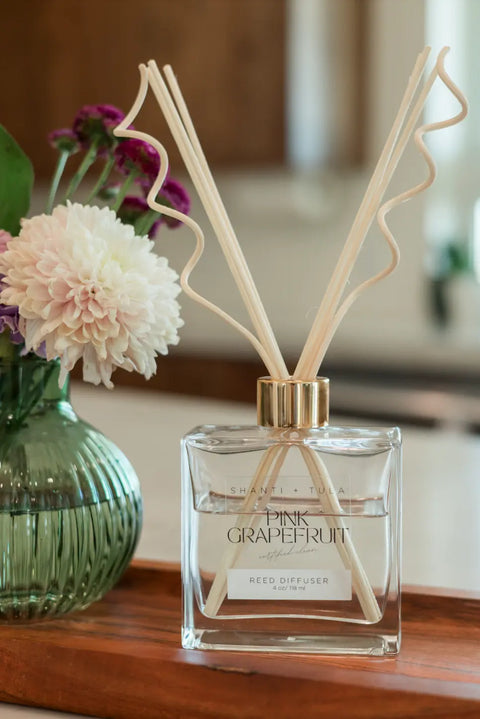A Shanti + Tula Flame-Free Reed Diffuser – Long-Lasting Aromatherapy for Home or Office sits on a wooden tray beside a green vase of white and purple flowers, adding refreshing fragrance and a lovely touch to your space. Made in the USA.