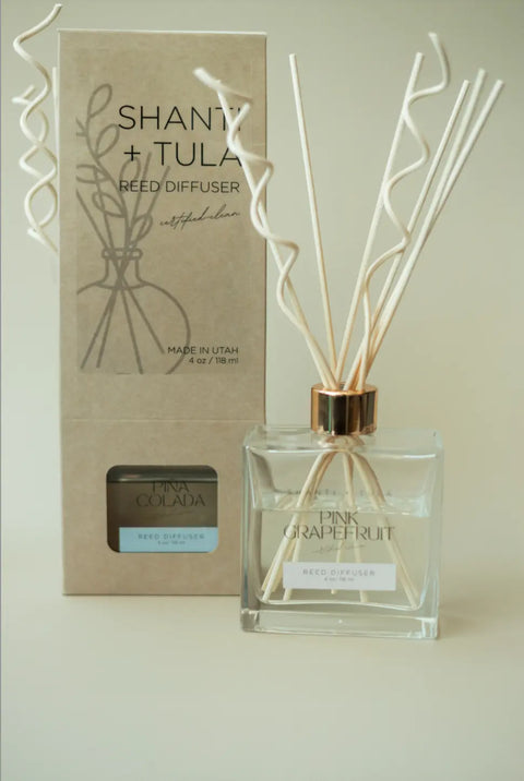 A clear Flame-Free Reed Diffuser from Shanti + Tula, labeled Pina Grapefruit with tan reeds, sits before a beige box—both featuring minimalist designs. Enjoy long-lasting aromatherapy for home or office, made in the USA.