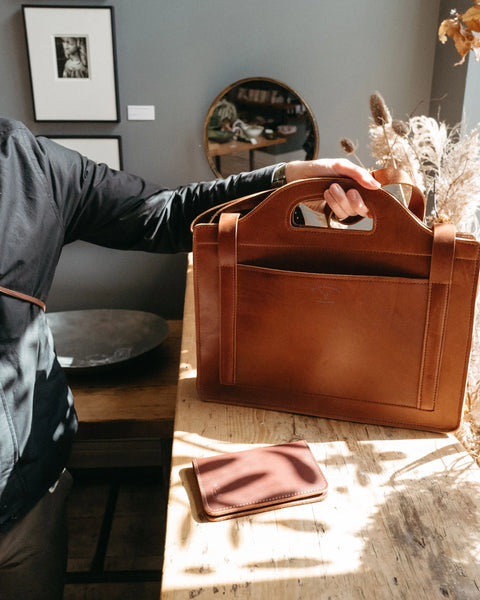 A person holds the Urban Southern Franklin Satchel, a brown professional bag, on a wooden table beside a matching leather wallet. Sunlight filters in through a window, with framed art and a round mirror visible in the background.