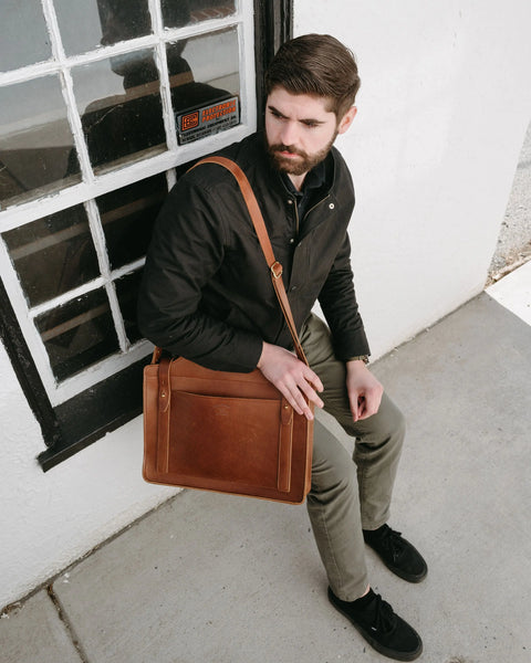 A man with brown hair and a beard, dressed in a black jacket and khaki pants, sits by a window ledge holding the Urban Southern Franklin Satchel laptop bag, gazing thoughtfully to the side.
