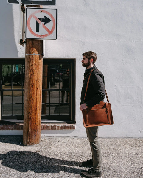 A man with a brown Urban Southern Franklin Satchel stands on a sidewalk next to a wooden pole and a no right turn sign, with a white building and street-reflecting window in the background.