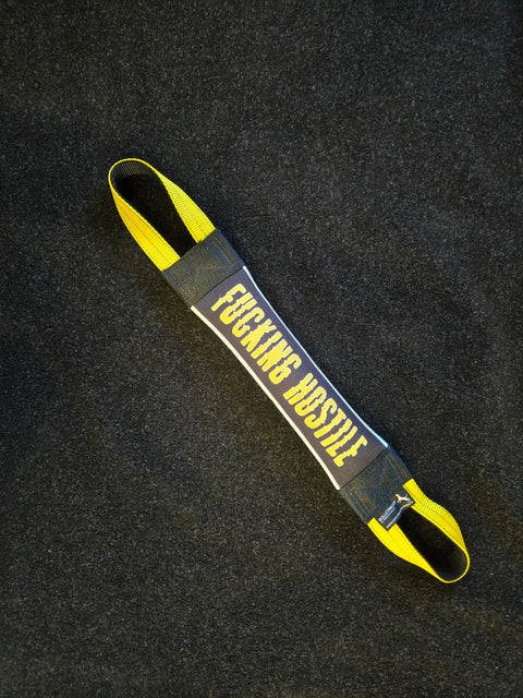 The Fucking Hostile Fire Hose Training Tug (Bad Tug Line) by Bulletproof Pet Products Inc, a black and yellow heavy-duty dog toy styled like an exercise band, lies on a dark textured surface with FUCKING HUSTLE in bold yellow letters.
