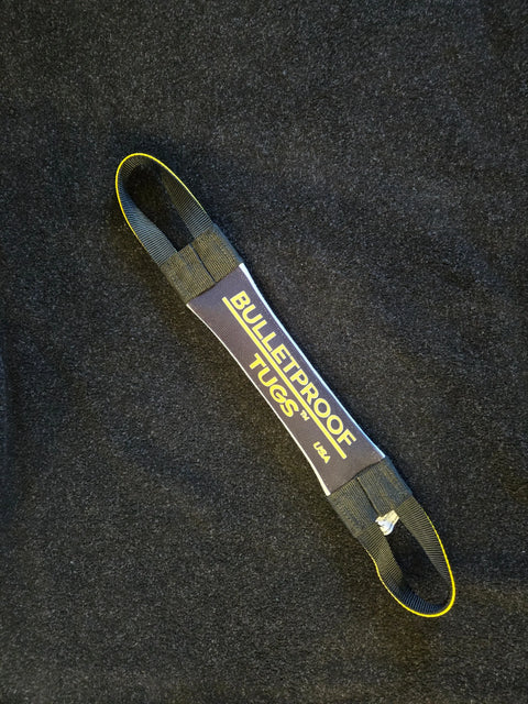 A black and yellow Fucking Hostile Fire Hose Training Tug (Bad Tug Line) from Bulletproof Pet Products Inc lies on a dark textured surface. This heavy-duty dog tug toy has two loops on each end.