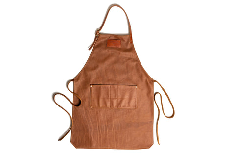 The Urban Southern Leather Apron features brown leather, an adjustable neck strap, a large front pocket, and side waist ties—ideal for grilling or daily tasks. Shown laid flat on a white background.