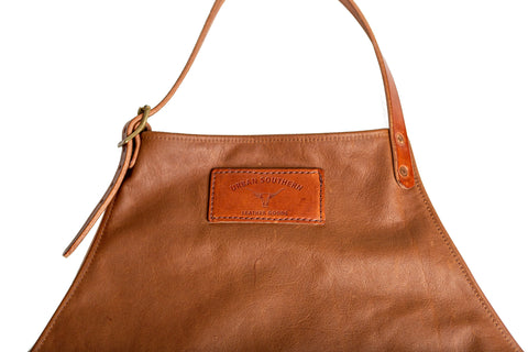 A close-up of a brown leather handbag with a brass buckle and a stitched Urban Southern label on the front, set against a white background—crafted with the same care as Urban Southern’s Leather Apron.