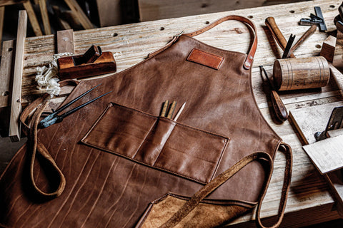 An Urban Southern Leather Apron rests on a wooden workbench, its pockets filled with tools, surrounded by woodworking equipment like a mallet, chisel, plane, and ruler in a busy workshop.