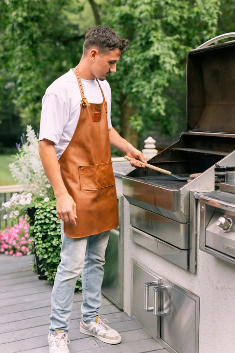 A young man wearing the Urban Southern Leather Apron stands on a deck grilling food outdoors on a stainless steel barbecue, holding a grilling tool and surrounded by greenery and potted flowers.