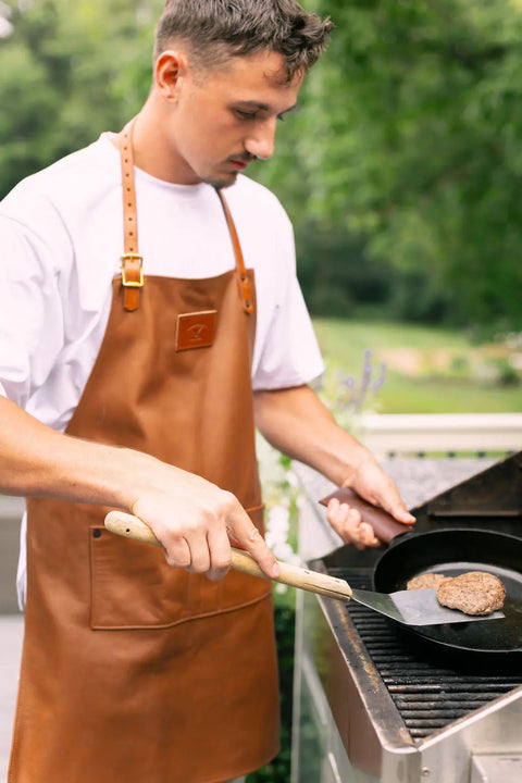 A man wearing an Urban Southern Leather Apron and a white T-shirt cooks a burger patty in a skillet on an outdoor grill with a spatula, with green trees and a yard visible in the background.
