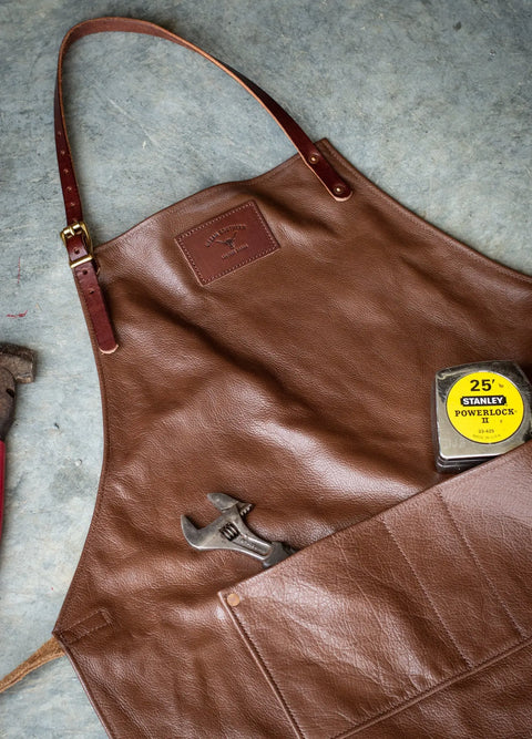An Urban Southern Leather Apron with front pockets holds a wrench and a 25-ft Stanley PowerLock tape measure. A tool handle is visible next to the apron on a gray surface.