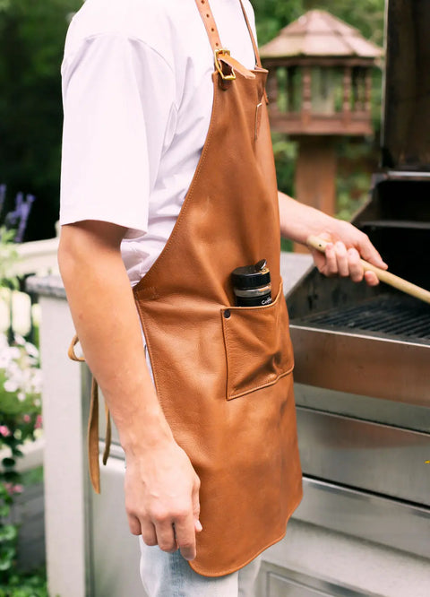 Wearing an Urban Southern Leather Apron and a white t-shirt, a person stands by an outdoor grill holding a grilling tool. A spice container is tucked in the apron pocket, with a wooden birdhouse in the background.
