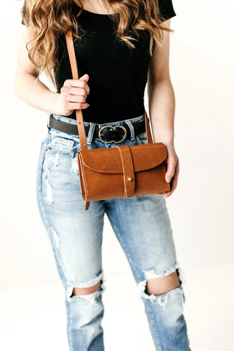 A woman in a black top and ripped blue jeans holds the Urban Southern Crossbody Wallet—a brown leather accessory ideal for minimalist style. The image is cropped at her shoulders, highlighting her wavy brown hair.