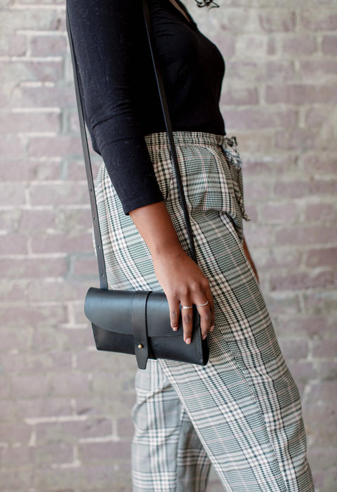 Wearing a black long-sleeve shirt and plaid pants, a person stands sideways by a brick wall, holding the Urban Southern Crossbody Wallet—a sleek accessory that adds minimalist style. Only their torso and upper legs are shown.