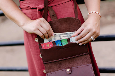 A person in a pink dress places cash and cards into the Urban Southern Crossbody Wallet, highlighting its minimalist brown leather design while wearing a bracelet and ring on their left hand.