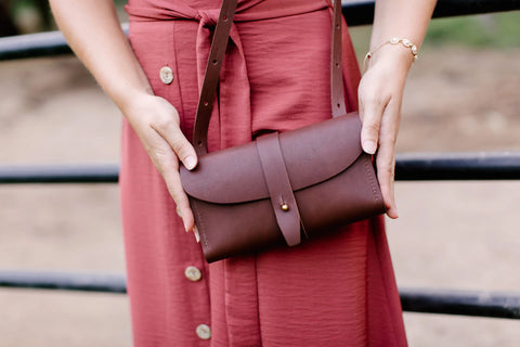 Wearing a rose-colored dress, a person holds the Urban Southern Crossbody Wallet, a dark brown leather accessory with strap and button closure, showcasing minimalist style while standing by a black metal fence.