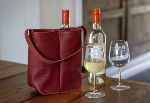 The Urban Southern Urban Dual Wine Tote, a red full-grain leather carrier holding two bottles of white wine, sits on a wooden table beside two filled wine glasses and an extra wine bottle in the background.