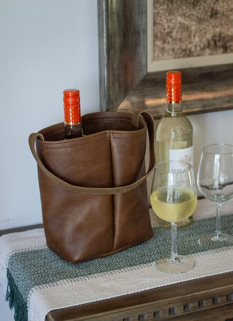 An Urban Southern Urban Dual Wine Tote, holding a wine bottle, sits on a table beside another wine bottle and two filled wine glasses. A framed picture hangs on the wall in the background.