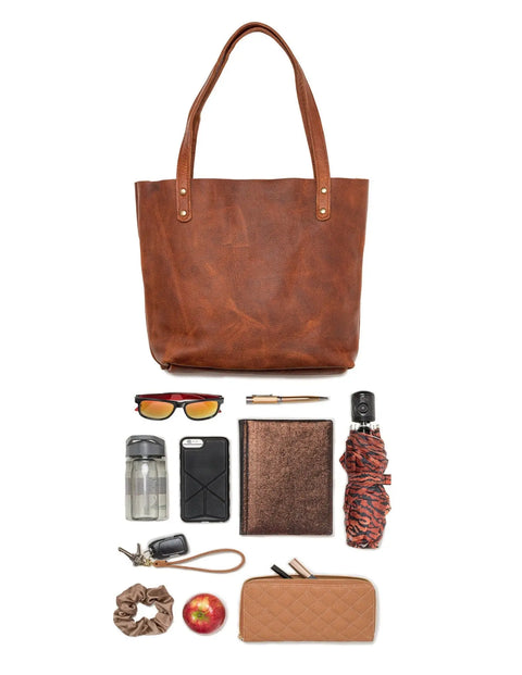 A Rogue Industries Fore Street Tote Bag sits above sunglasses, a pen, car keys, a water bottle, two phones, a brown notebook, colorful umbrella, scrunchie, red apple, and tan wallet—all neatly arranged on a white background.
