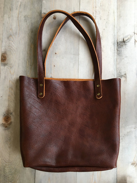 The Fore Street Tote Bag by Rogue Industries, crafted in Maine from brown full-grain leather, rests on a light wood surface. Its design is simple and unadorned, with visible stitching and metal rivets securing the handles.