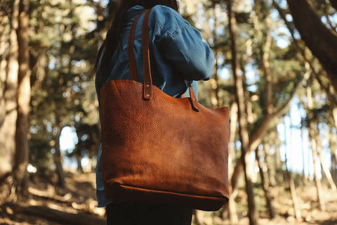 A person outdoors among tall trees carries a Rogue Industries Fore Street Tote Bag on their shoulder, paired with a blue denim shirt.